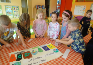 Szóstka dzieci stoi wokół stolika na którym leży plakat ekologiczny, dwóch chłopców nakleja elementy z kolorowego papieru.